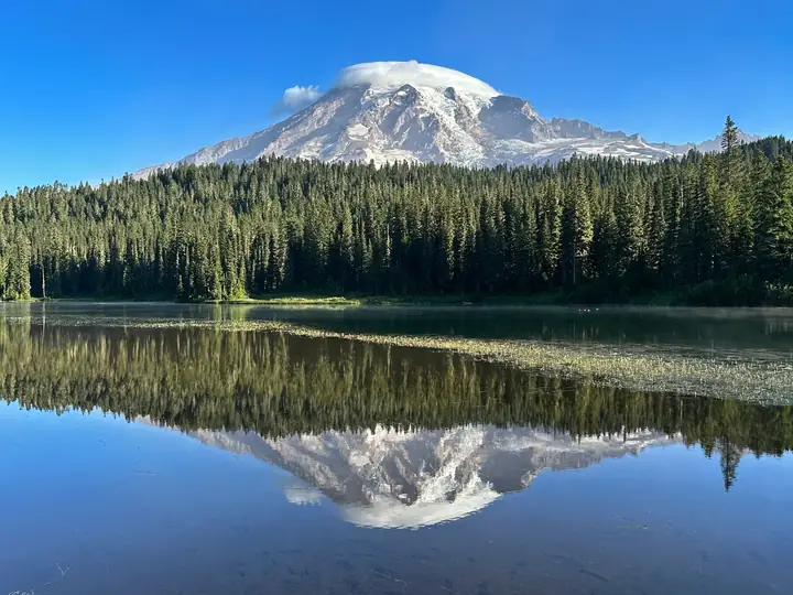Lago de montanha com floresta de pinheiros e um pico nevado refletidos na água parada
