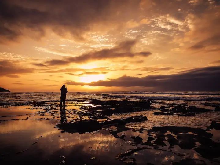 Pescador de cana em silhueta numa costa rochosa com o céu alaranjado do pôr do sol